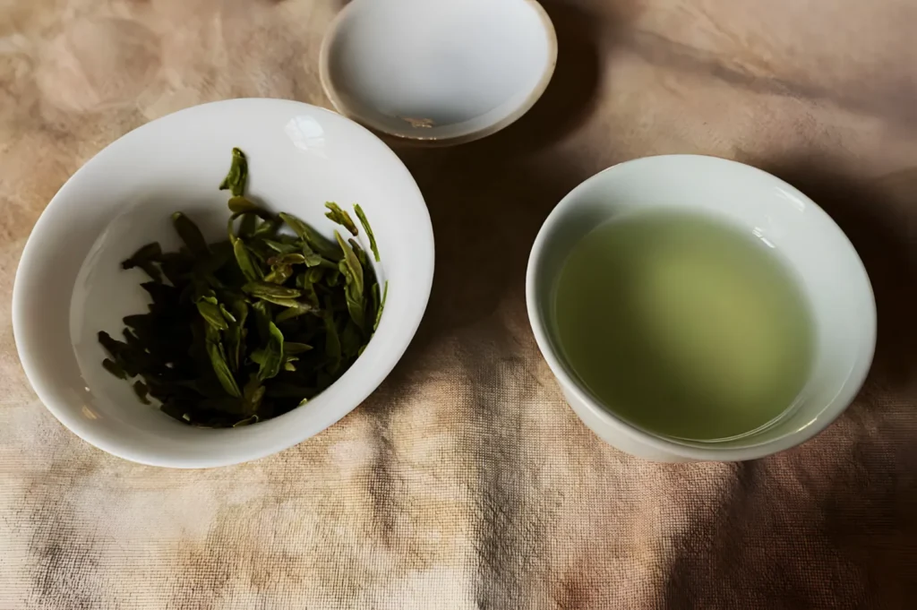 White bowls on a textured cloth; the left bowl holds fresh green tea leaves, while the right contains light golden brewed tea, evoking a serene, calming vibe.