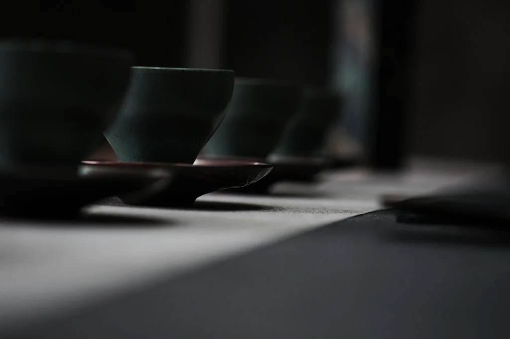 Close-up of green ceramic cups on matching saucers, arranged in a line on a table. The scene has a serene and minimalist atmosphere.