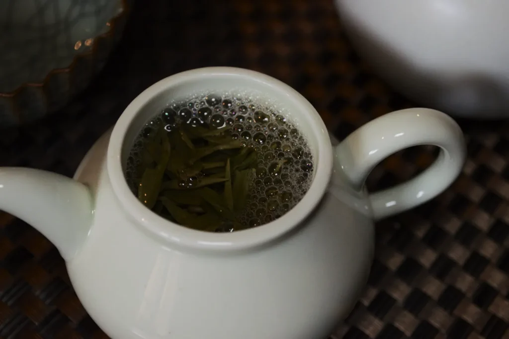 A small white teapot filled with green tea and visible tea leaves, surrounded by bubbles, sits on a dark woven mat, creating a cozy, inviting atmosphere.