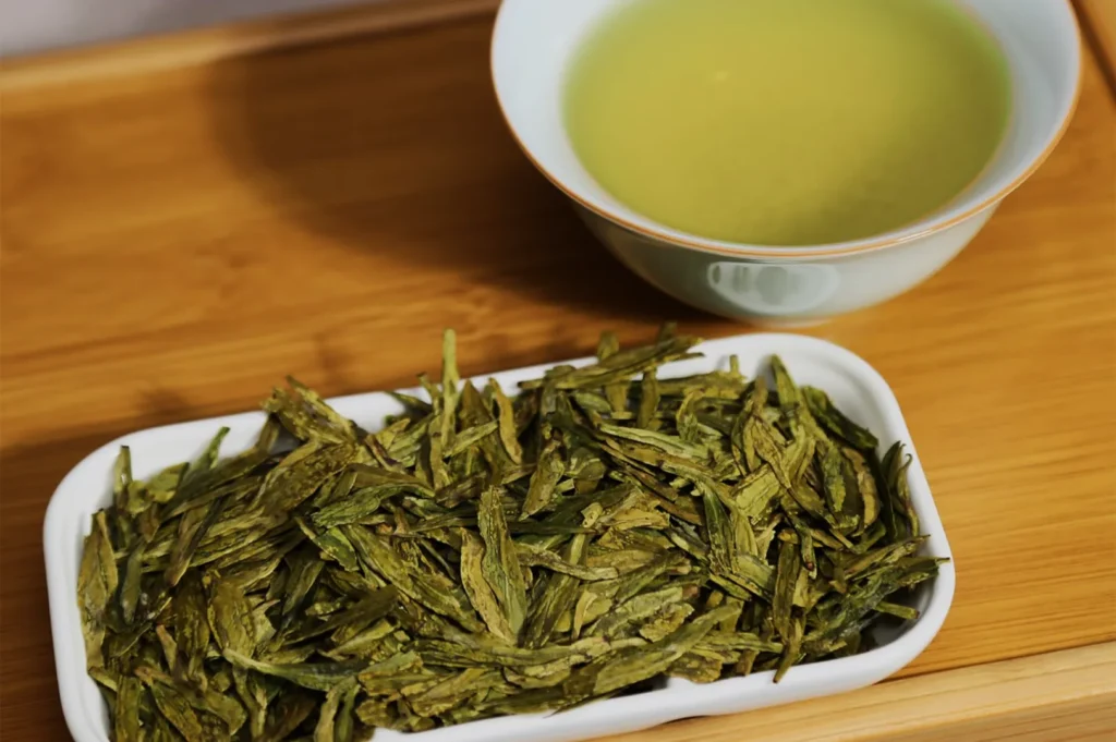 A wooden surface displays a bowl of light green tea and a rectangular dish filled with dried green tea leaves, conveying a sense of freshness and calm.