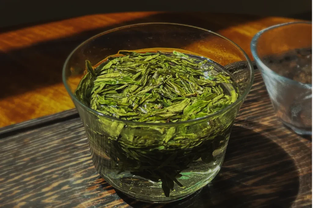 A glass cup filled with vibrant green tea leaves soaking in water sits on a wooden surface, highlighted by warm sunlight, creating a calming ambiance.