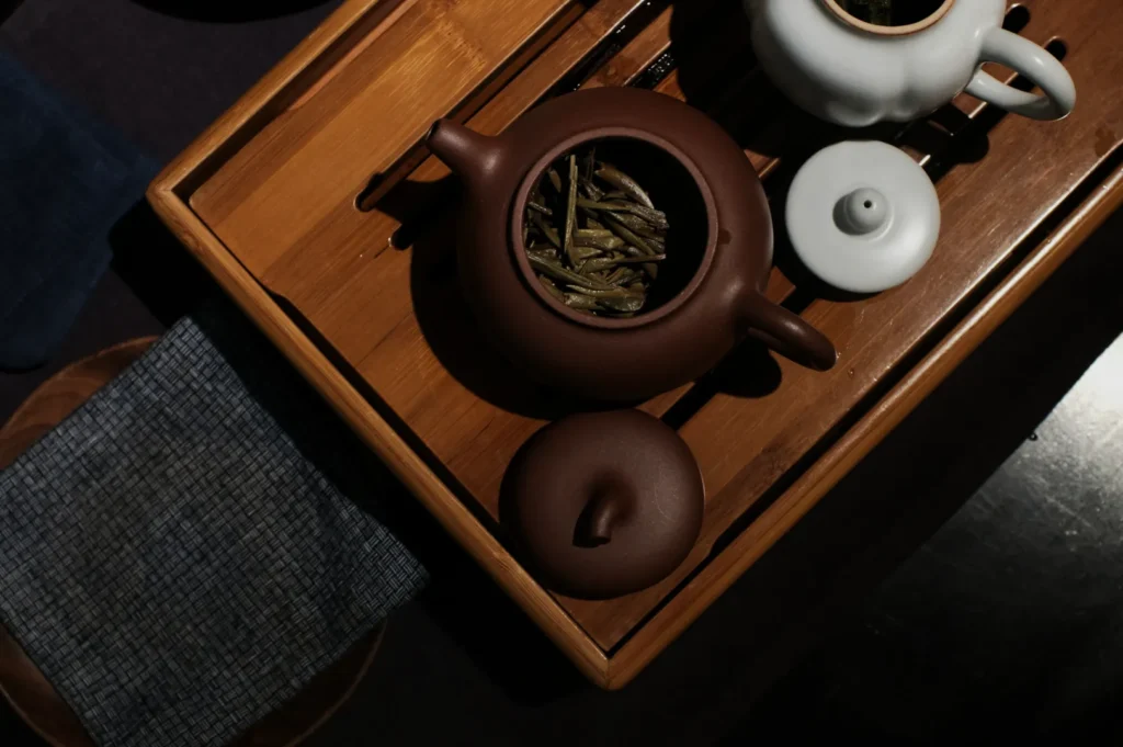 Overhead view of a brown teapot with green tea leaves, a white teapot, and a wooden tray, creating a calm, elegant tea setting.