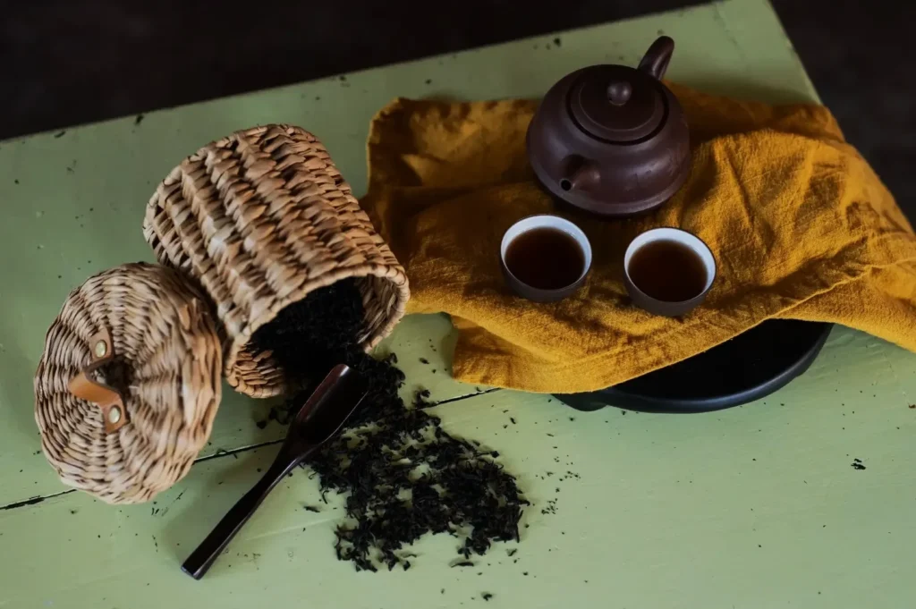 Wicker basket with loose tea, wooden scoop, brown teapot, and two tea cups on yellow cloth on light green surface, evokes a cozy atmosphere.