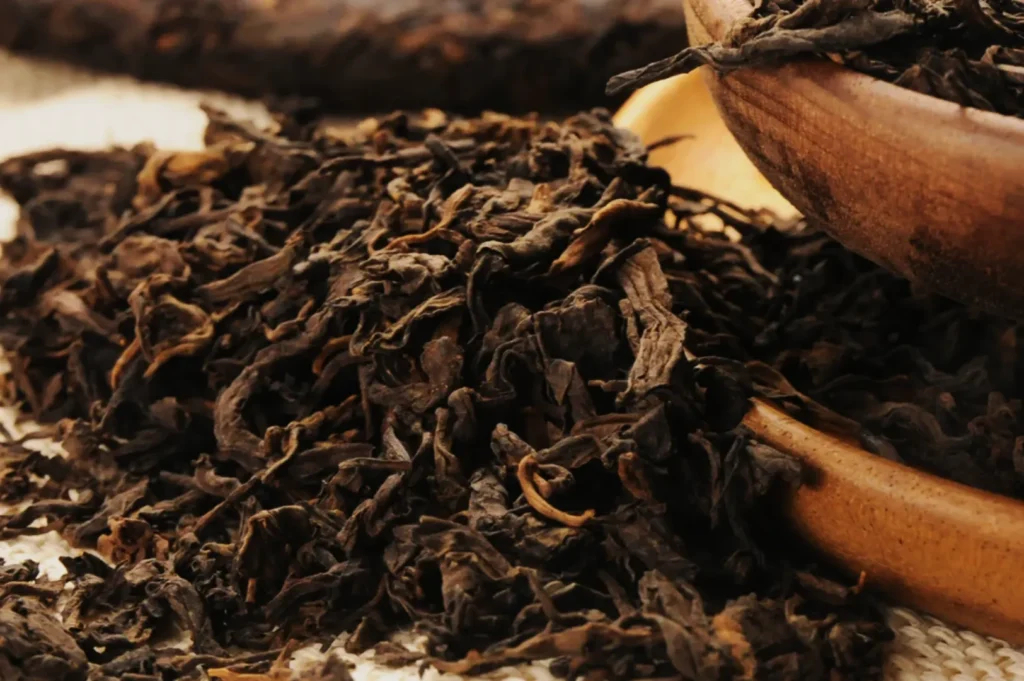Close-up of dried Pu-erh tea leaves spilling from a wooden bowl onto a textured surface. The rich, dark brown leaves convey an earthy tone.