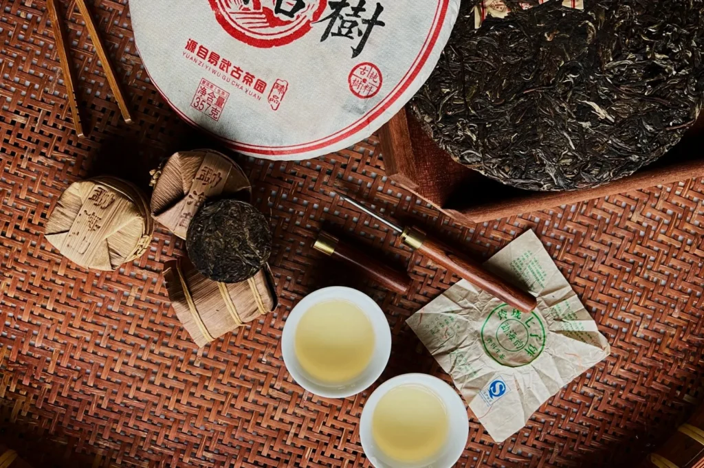 Top-view of a bamboo tray with compressed tea cakes, a wrapped tea package, two wooden tea picks, and two cups of light-colored tea, conveying a traditional and calming ambiance.