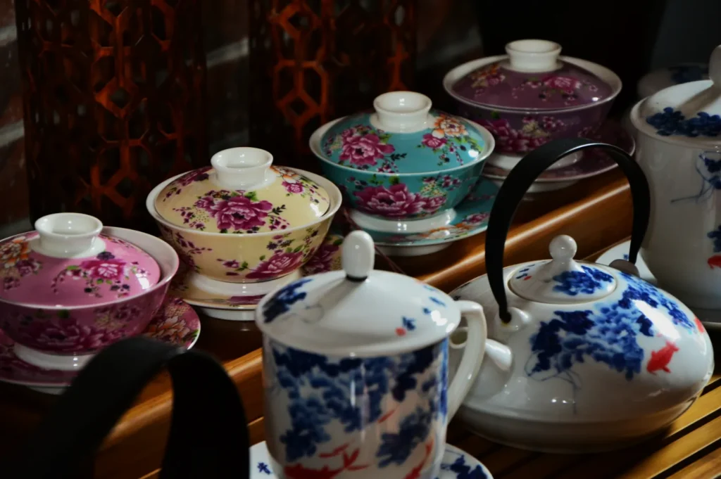 A collection of ornate teacups and teapots with floral patterns in pink, blue, and purple hues, arranged on a wooden tray with a warm, elegant tone.