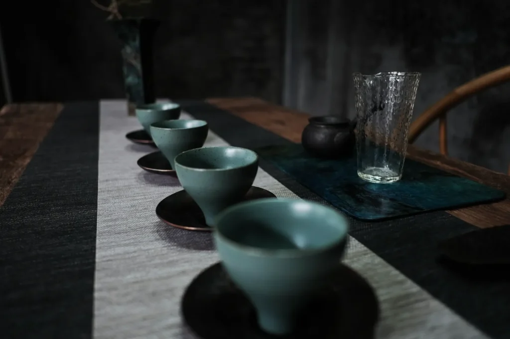 A row of teal ceramic cups on dark saucers sits on a striped table runner. A clear glass and small teapot rest on a teal placemat, creating a serene tea setting.