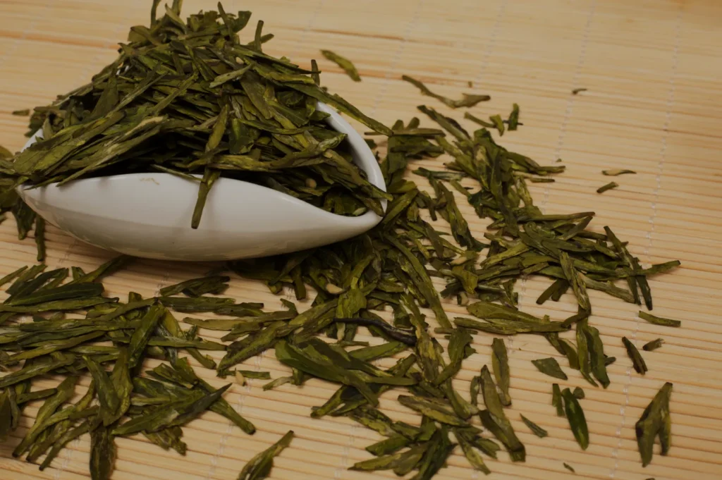 Loose green tea leaves spill from a white bowl onto a bamboo mat. The vibrant green color suggests freshness, evoking a sense of natural serenity.