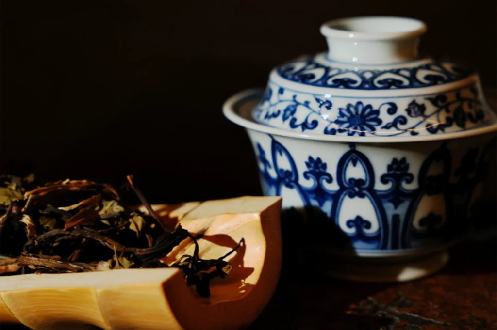 A blue and white porcelain cup with intricate floral patterns is next to a wooden tray holding dried tea leaves, evoking a traditional, serene ambiance.