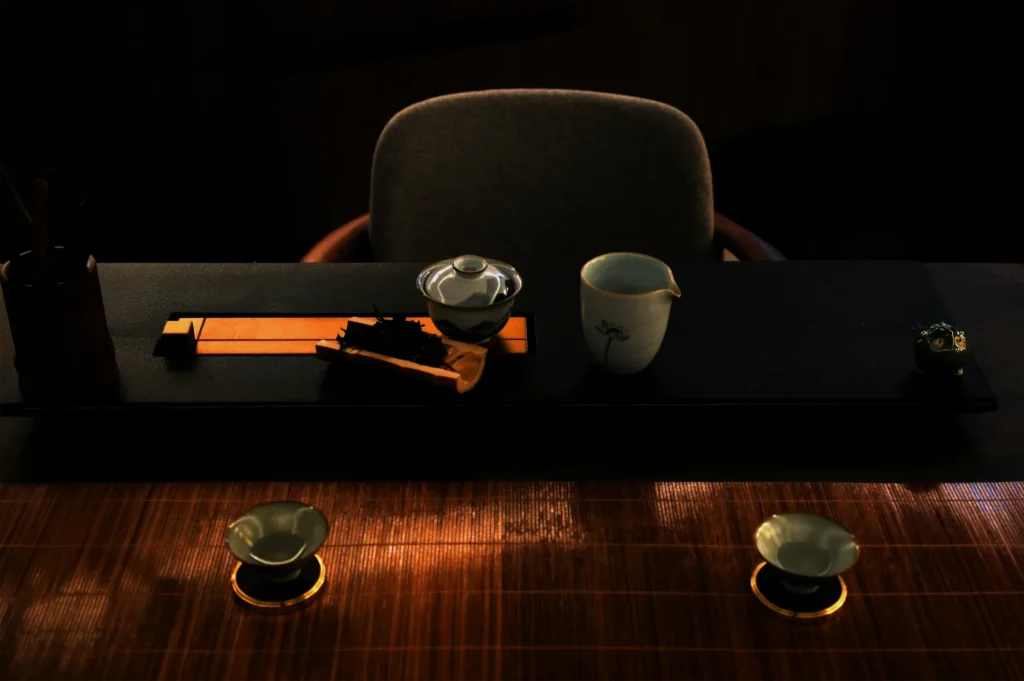 Dimly lit tea setup with a teapot, cups, and accessories on a dark tray. The scene exudes calm and tradition, with warm wood tones and soft shadows.