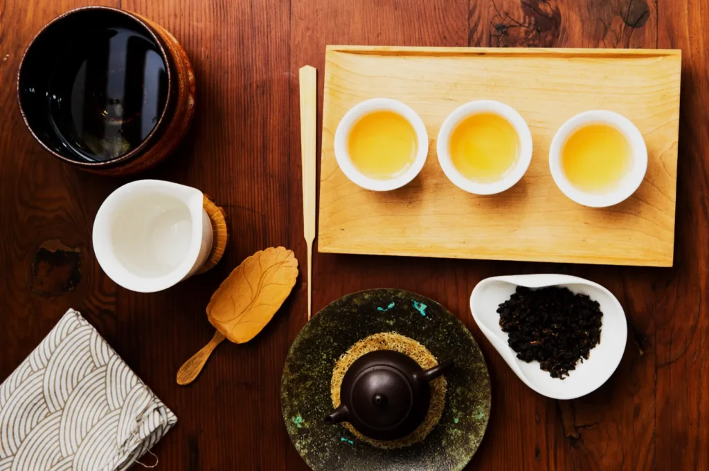 A top-down view of a tea set on a wooden table with three cups of amber tea on a tray, a teapot, tea leaves in a dish, and a leaf-shaped spoon.