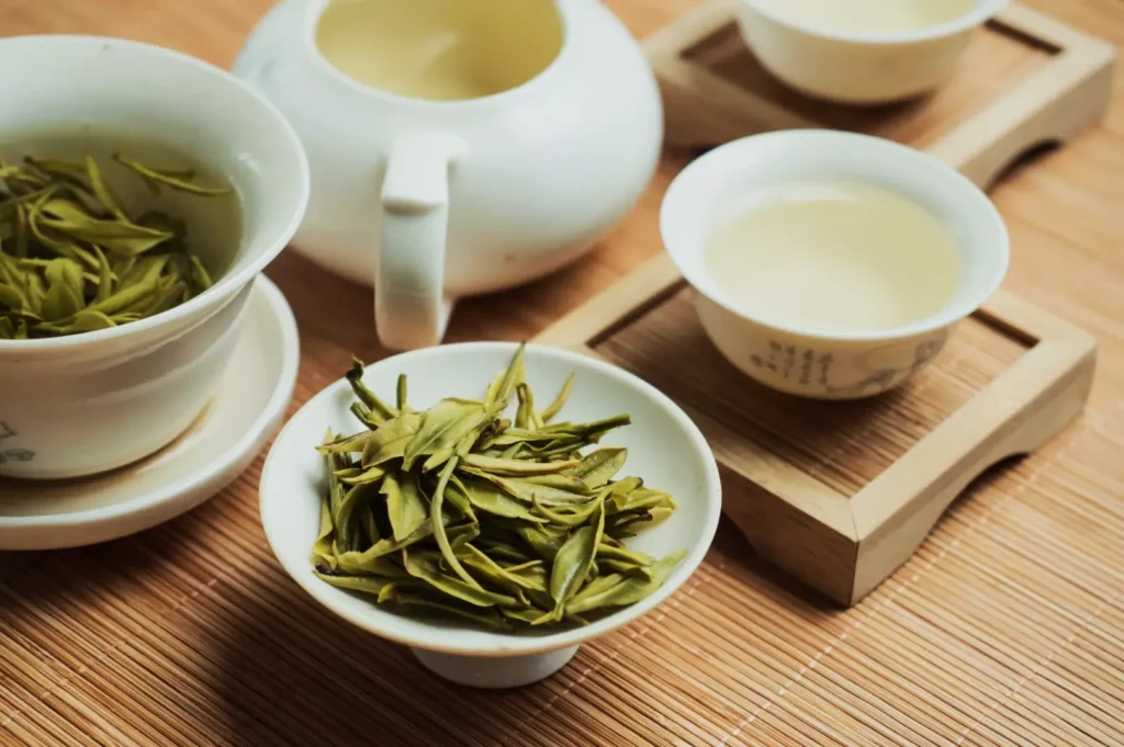 White teapot and cups with yellow tea, dried green leaves in bowls, set on a bamboo mat. The ambiance is warm and inviting.