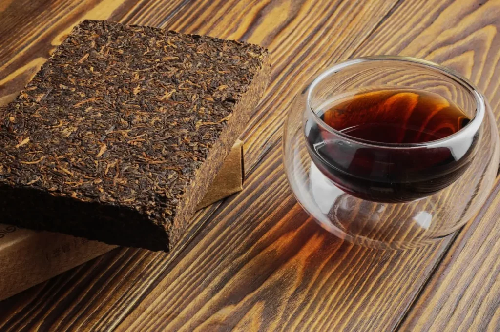 A compressed block of dark tea and a glass cup of brewed tea sit on a wooden table. The rich tea contrasts with the warm, textured wood surface.