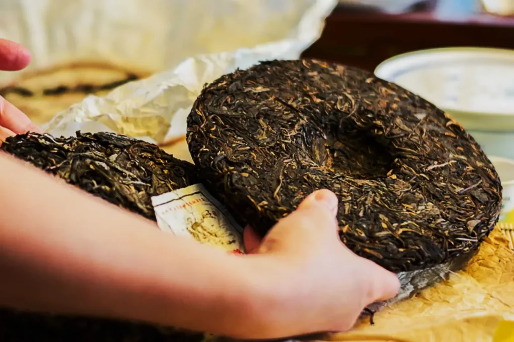 Hands holding a round, pressed tea cake wrapped in paper. The textured surface shows dark tea leaves, indicating traditional tea preparation.
