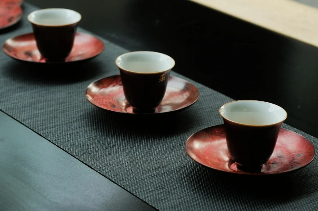 Three ceramic cups with white interiors sit on red saucers, lined up on a textured, dark table runner. The arrangement feels calm and orderly.