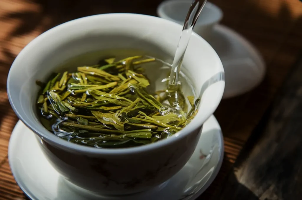 White teacup with fresh green tea leaves being steeped in hot water. Minimalistic and serene, with soft lighting and a wooden surface.