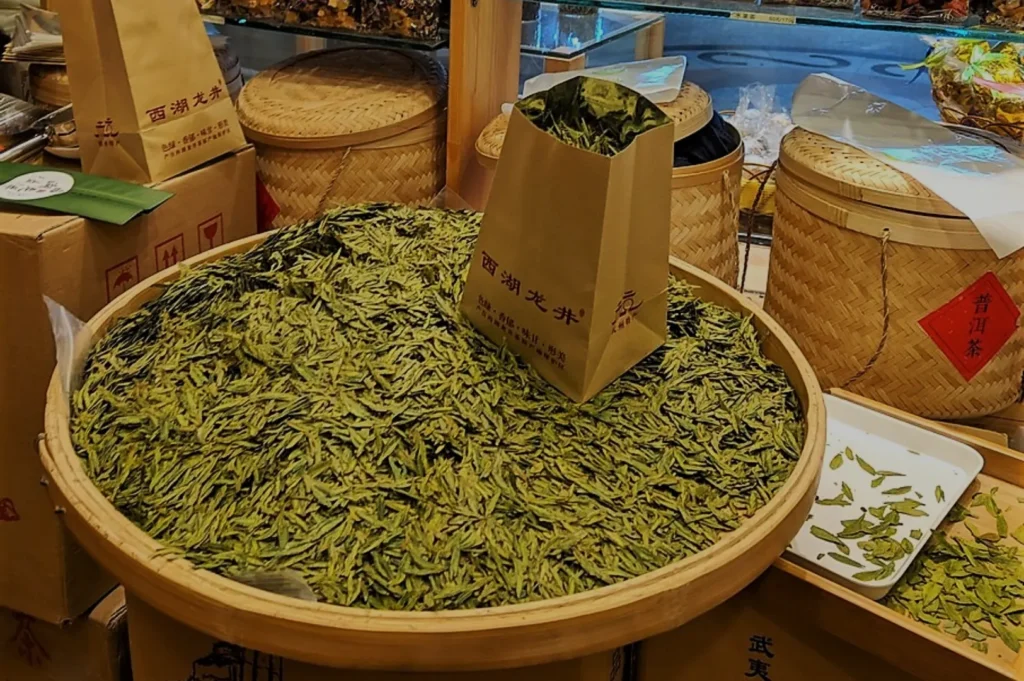 A large bamboo basket filled with loose green tea leaves, with a brown paper bag placed on top. Nearby are wicker baskets and cardboard boxes.