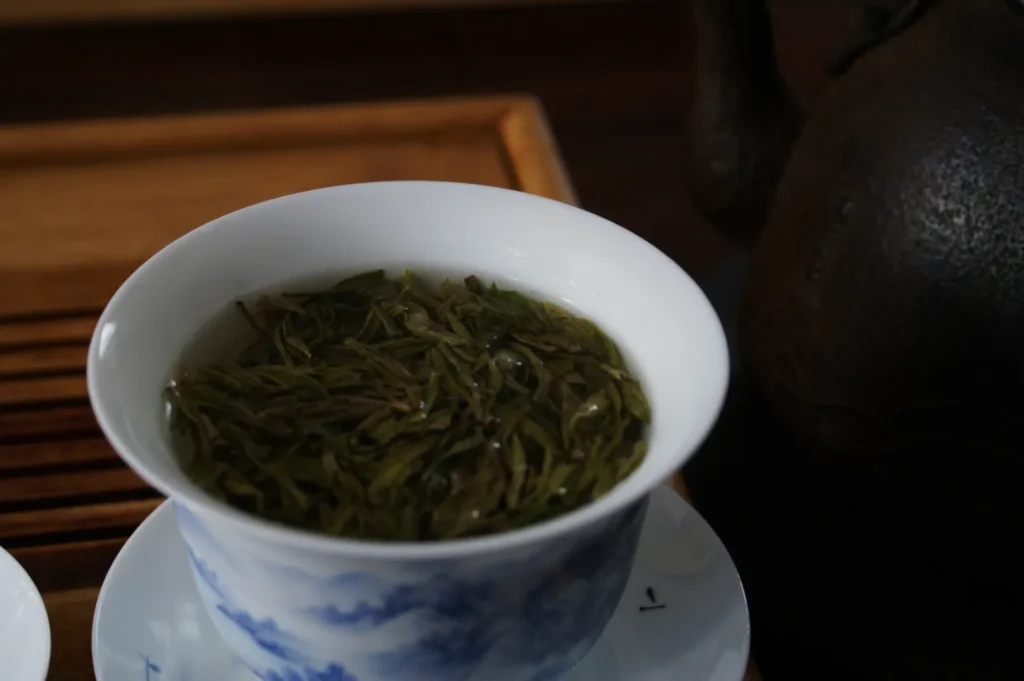A white teacup with blue patterns holds loose green tea leaves steeping in water. The setup rests on a wooden tray, near a dark teapot, evoking calmness.
