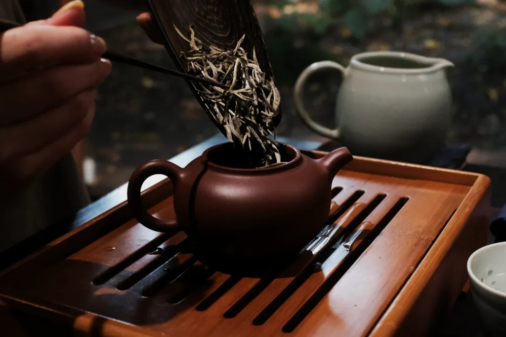 A hand pours dried tea leaves from a wooden scoop into a small, brown clay teapot on a wooden tray. A serene atmosphere reflects a tea ceremony.