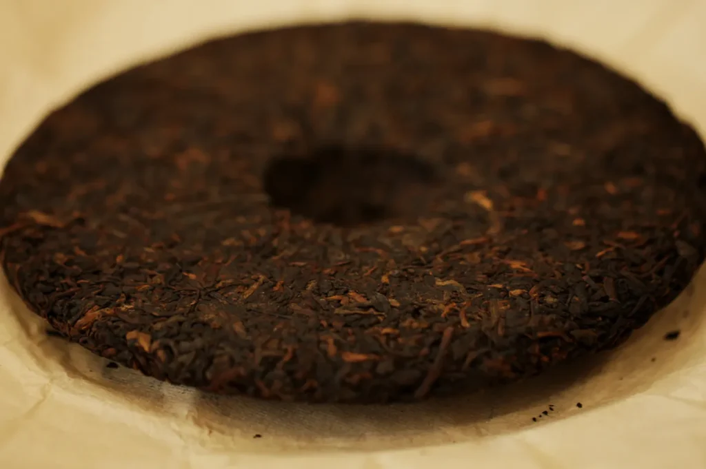 Round dark brown pu-erh tea cake with a hole in the center, resting on beige parchment. The surface is textured, conveying an earthy tone.
