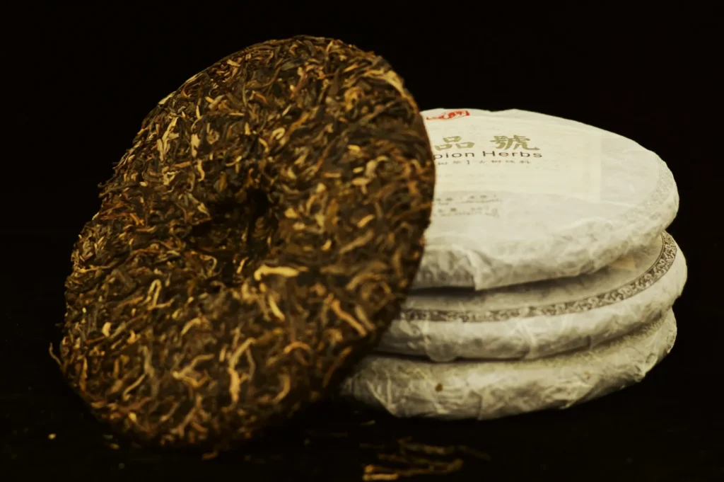 Tea cake with a dark, textured surface leans against a stack of wrapped, white tea cakes. The background is black, highlighting the natural tones.