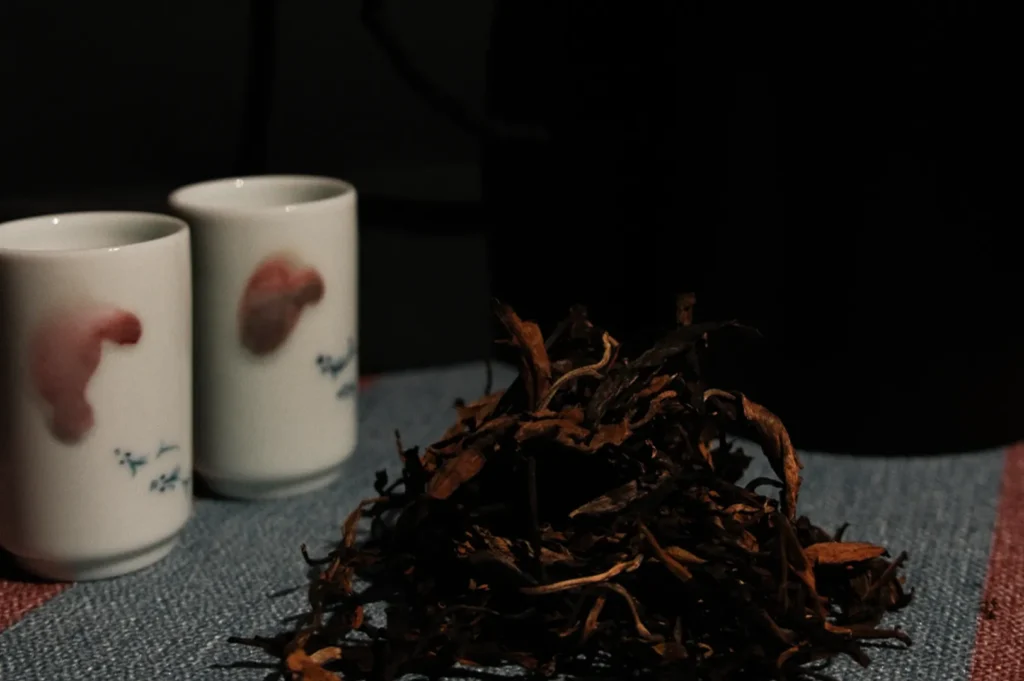 Two white cups with floral patterns stand on a textured cloth beside a small pile of loose tea leaves, creating a cozy and inviting atmosphere.