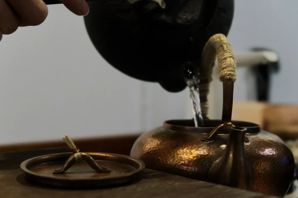 A hand pours water from a black kettle into a hammered copper teapot. The teapot lid is off, resting nearby. The tone is calm and focused.