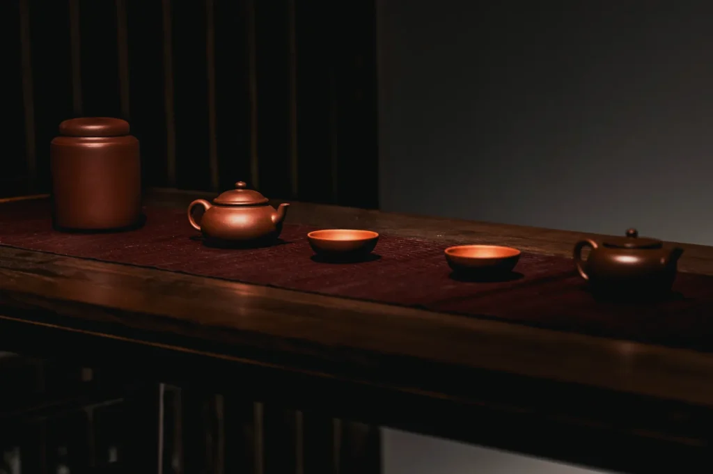 A set of traditional brown clay teaware, including a jar, teapots, and teacups, arranged on a wooden table with a maroon mat in a dimly lit setting.