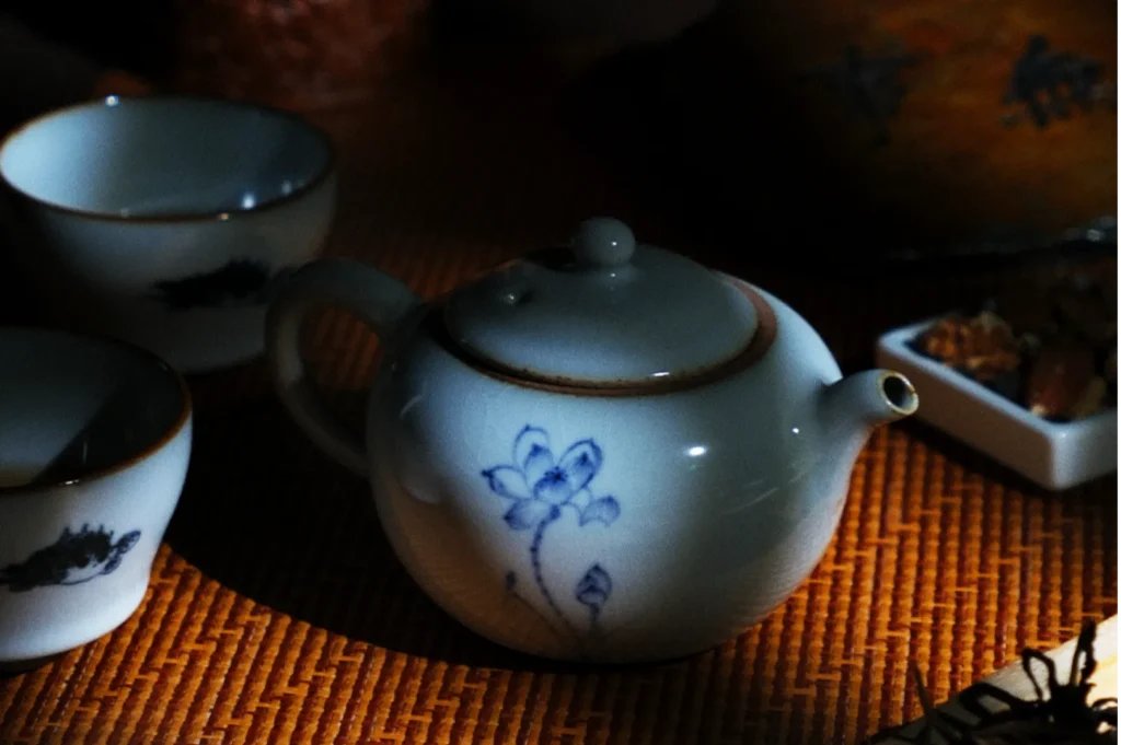 A small white teapot with blue floral designs sits on a woven mat, surrounded by two cups and a dish with nuts. The scene is warmly lit, evoking a cozy, tranquil atmosphere.