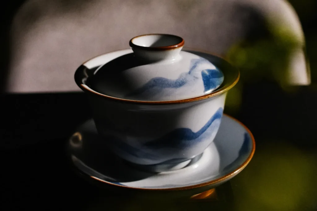 A ceramic tea cup with a lid, decorated with subtle blue brushstrokes and brown trim, sits gracefully in soft, moody lighting on a saucer.