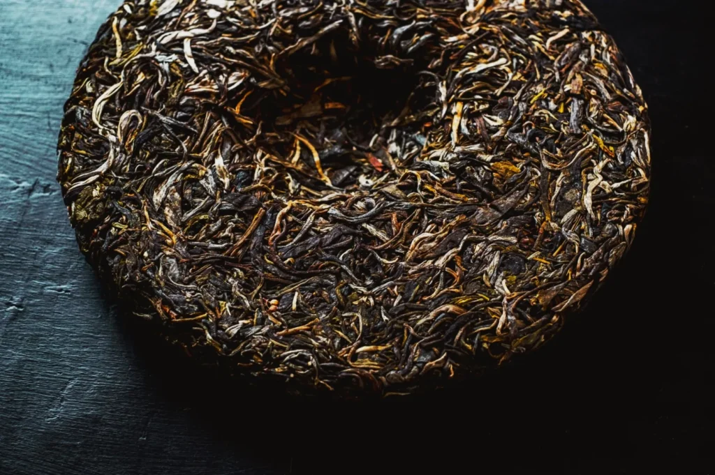 A round, compressed tea cake with tightly packed, dried tea leaves in shades of brown and green on a dark textured surface, conveying rustic simplicity.