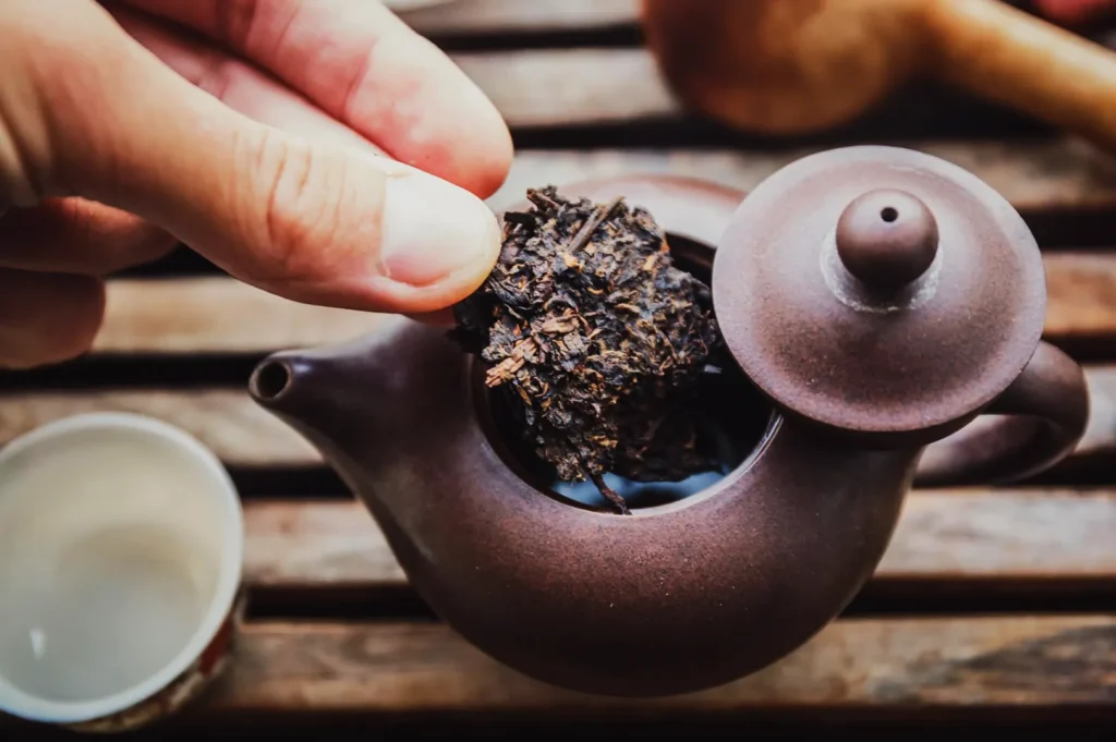 A hand places a chunk of dark tea leaves into a small, brown teapot on a wooden table. A nearby white cup suggests a serene tea preparation.