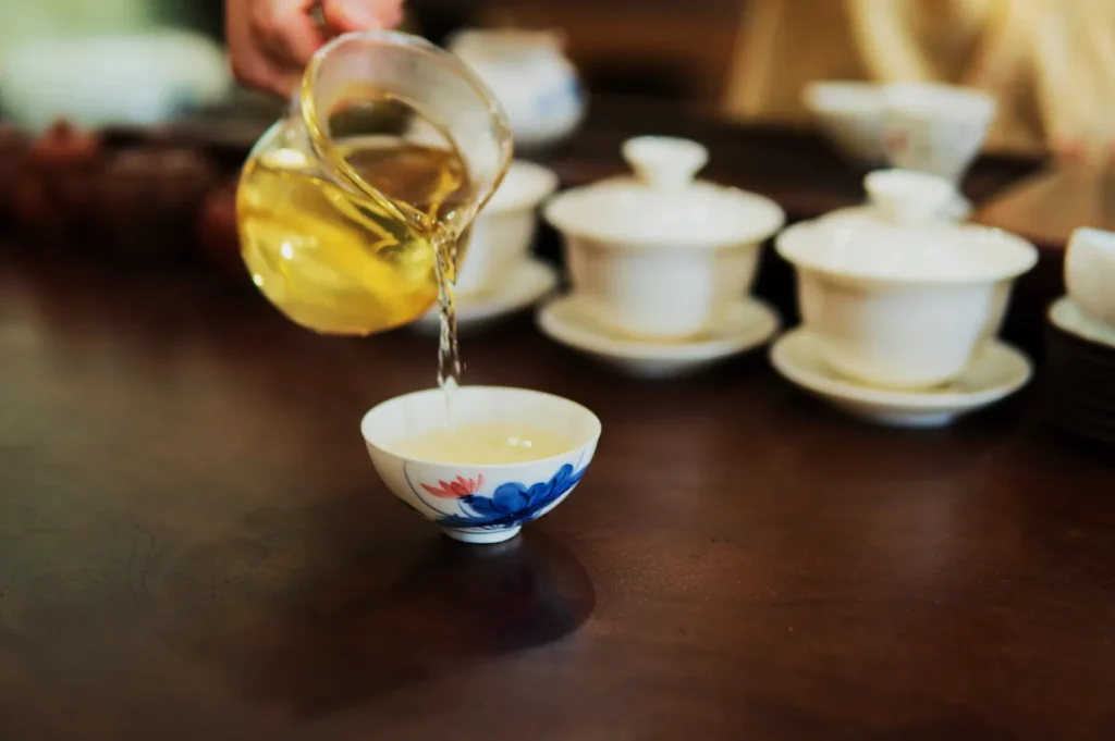 A hand pours golden tea from a glass pitcher into a floral teacup. White teacups with lids are in the background on a smooth brown table.