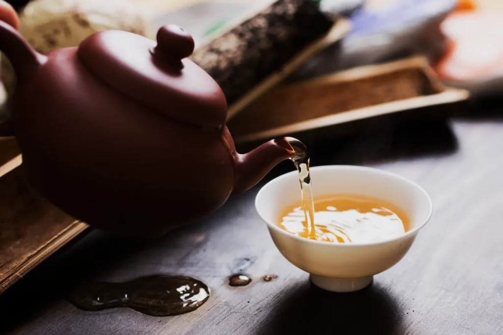 A brown teapot pours steaming tea into a small white cup on a dark wooden table, with spilled tea adding a cozy, inviting ambience.