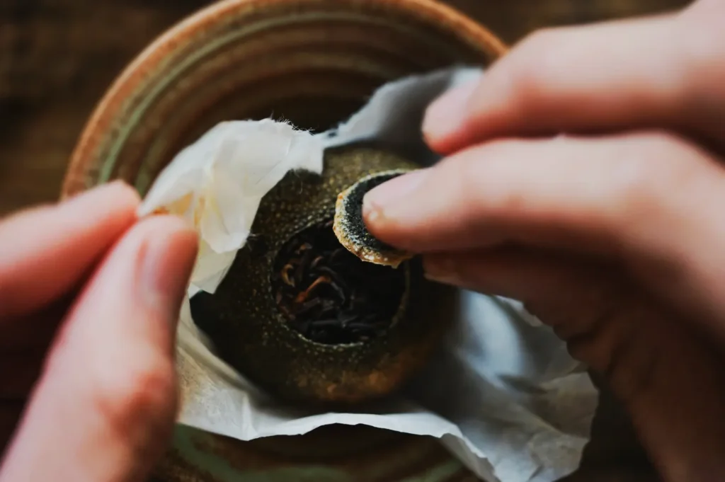 Hands gently peel a dried citrus peel in a bowl, revealing loose black tea leaves inside. The scene conveys a sense of care and artisanal craftsmanship.