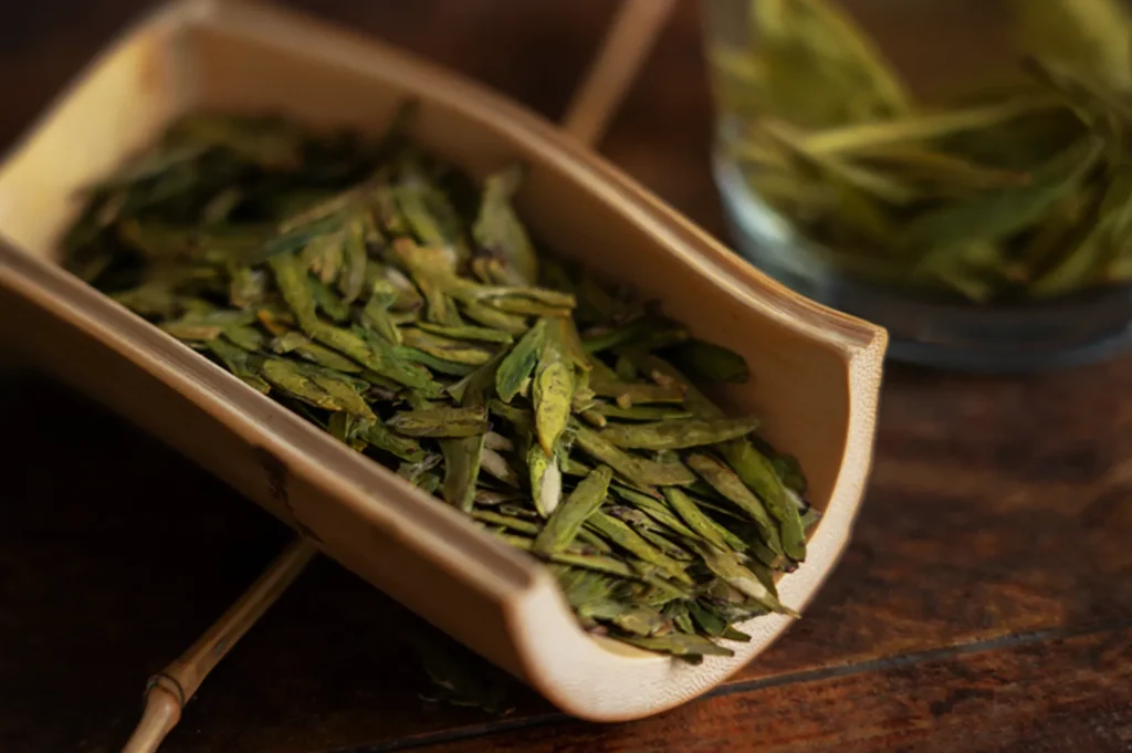 Loose leaf tea on a wooden scoop, displaying vibrant green colors, with a cup of brewed tea blurred in the background on a wooden table.