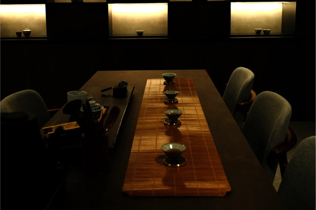 Dimly lit room with a wooden table set for tea, featuring five small teacups on a bamboo mat. Soft, ambient lighting creates a calm atmosphere.