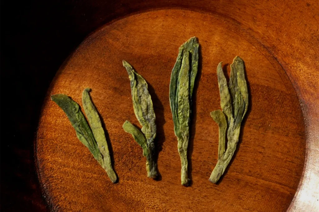 Four dried green tea leaves are arranged in a row on a wooden surface. The leaves have a slightly curled appearance and vary in size.