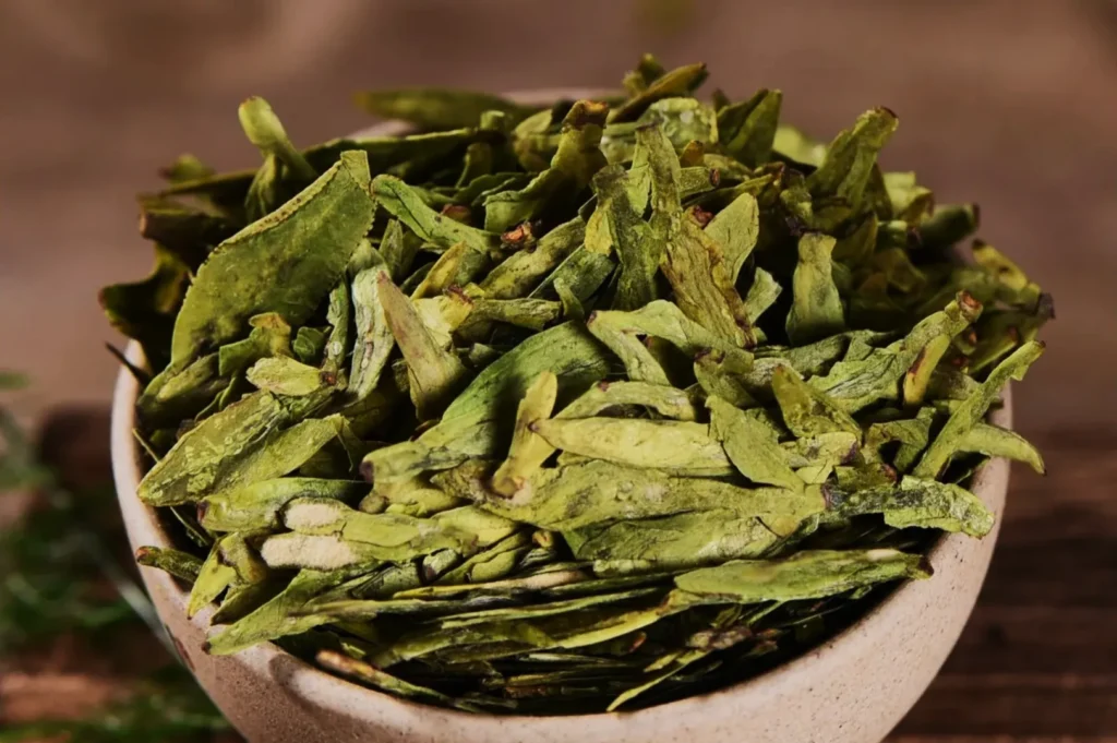 A bowl filled with dried green tea leaves sits on a wooden table, showcasing vibrant green hues and a rustic, natural setting.