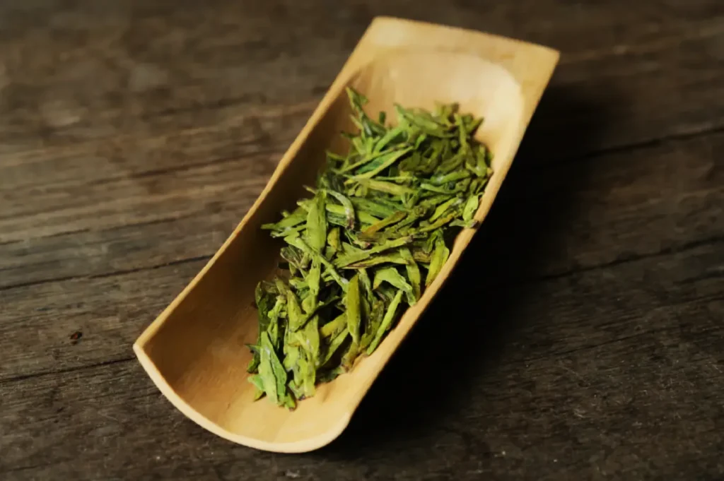 Dried green tea leaves in a wooden tray resting on a rustic wooden surface, conveying a natural and organic feel.