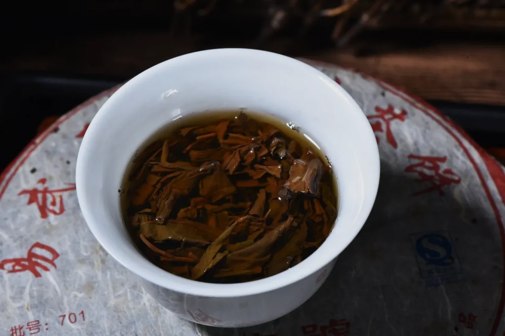 Close-up of a white cup filled with steeped tea leaves in brown liquid. The cup is on a textured surface with red Chinese characters, conveying a traditional tea experience.