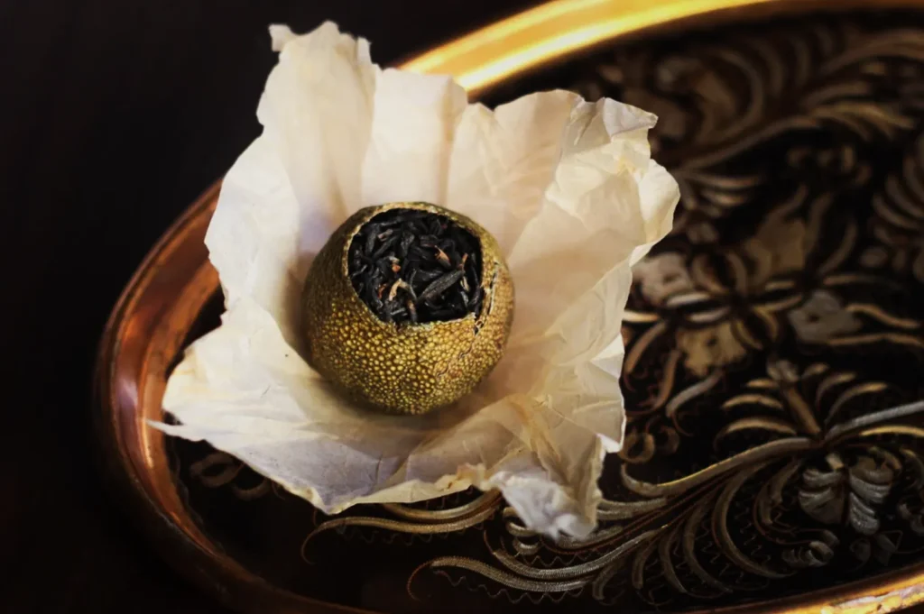A round citrus fruit filled with black tea sits open on crinkled white paper, placed on an ornate, dark wooden tray with gold floral patterns.