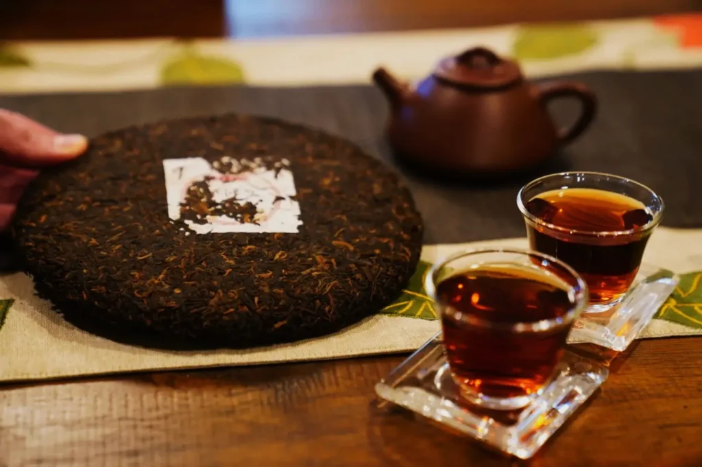 A hand holds a round cake of aged Pu-erh tea on a table. Nearby, a brown teapot and two steaming glass cups of tea sit, creating a cozy, inviting scene.