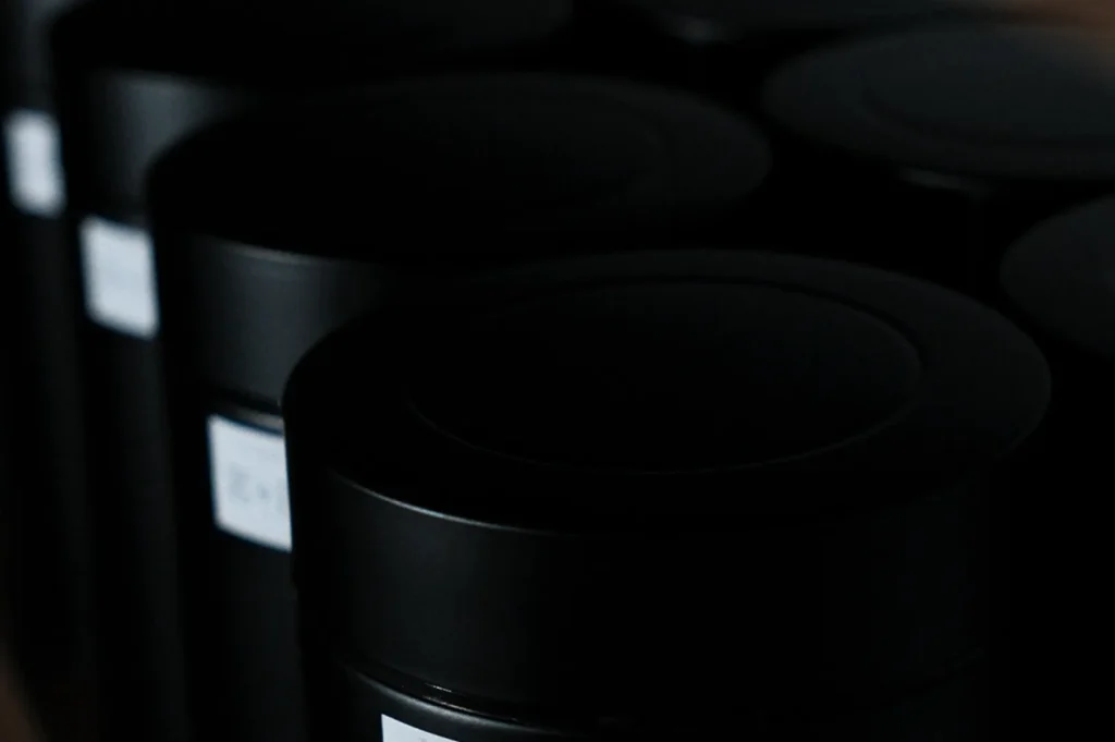 Close-up of black cylindrical containers with labels on a dark background. The image conveys a sleek, minimalistic, and mysterious tone.
