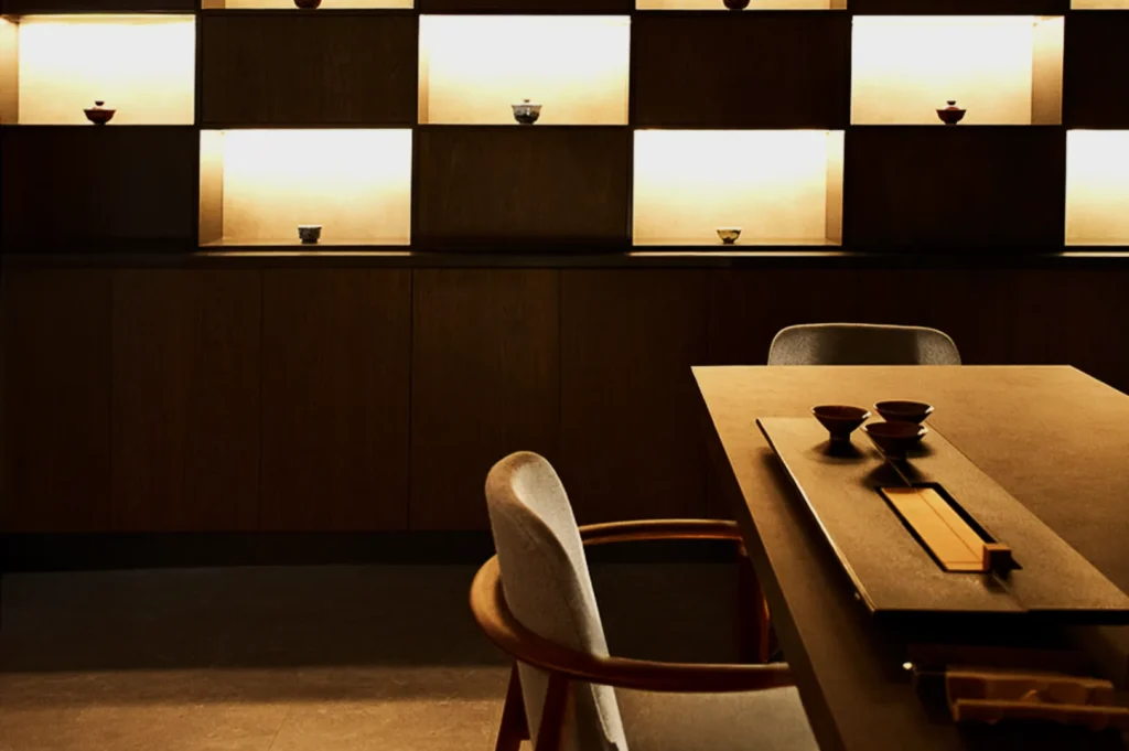 Minimalist room with a wooden table and soft chairs, featuring a tabletop tea set. Backlit shelves display small bowls, creating a serene ambiance.