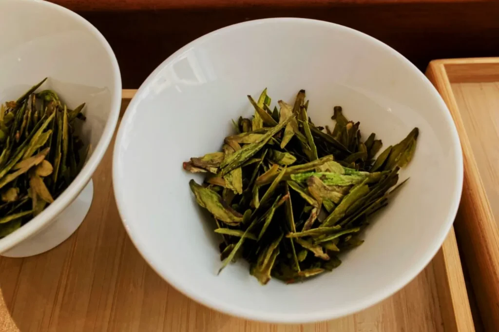 A white bowl on a wooden tray contains vibrant green tea leaves. The fresh, earthy tones of the leaves convey a sense of natural healthiness.