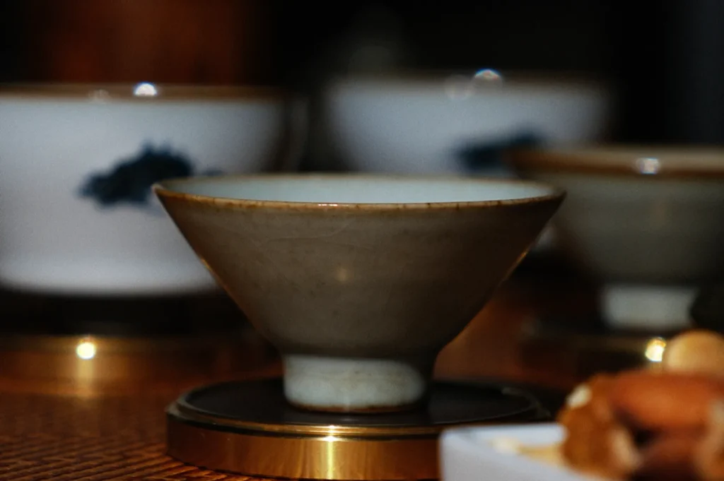 A close-up of a ceramic teacup with a subtle glaze, resting on a brass saucer. Blurred teacups in the background create a warm, elegant ambiance.