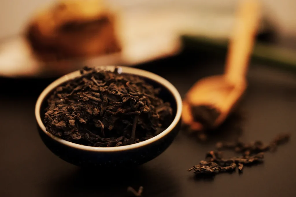 A close-up of loose leaf tea in a small blue bowl on a dark surface. A wooden scoop and more tea leaves are in soft focus in the background. The mood is warm and inviting.