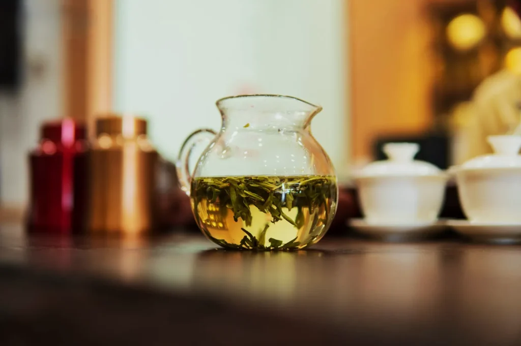 A clear glass teapot with floating green tea leaves sits on a wooden table. Blurred, colorful jars and white teacups create a warm, inviting ambiance.