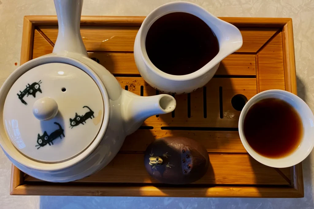 A bamboo tray holds a white teapot with black cat designs, a pitcher, a teacup with dark tea, and a small brown figurine, creating a warm, cozy tea scene.